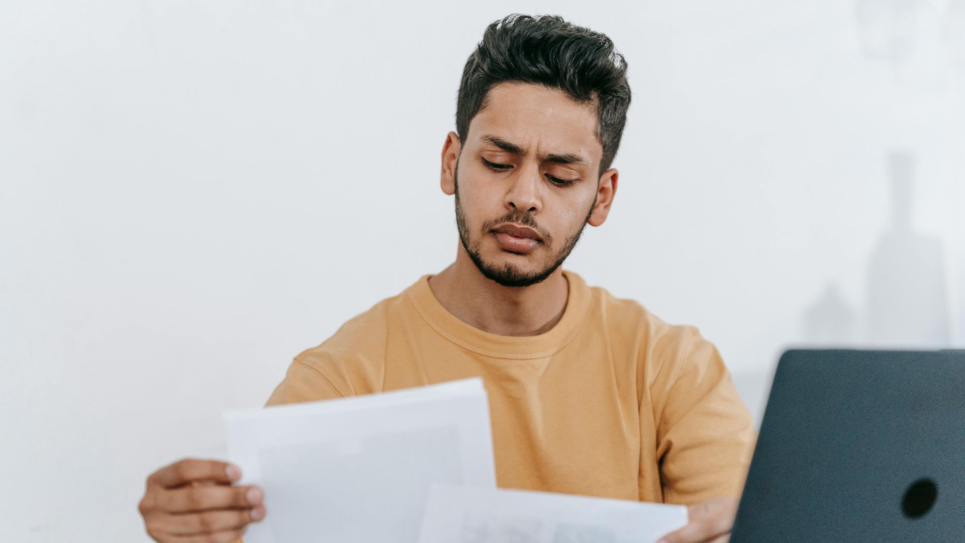 Focused young man reviewing paperwork at his desk, showcasing a business setting with a laptop indoors.