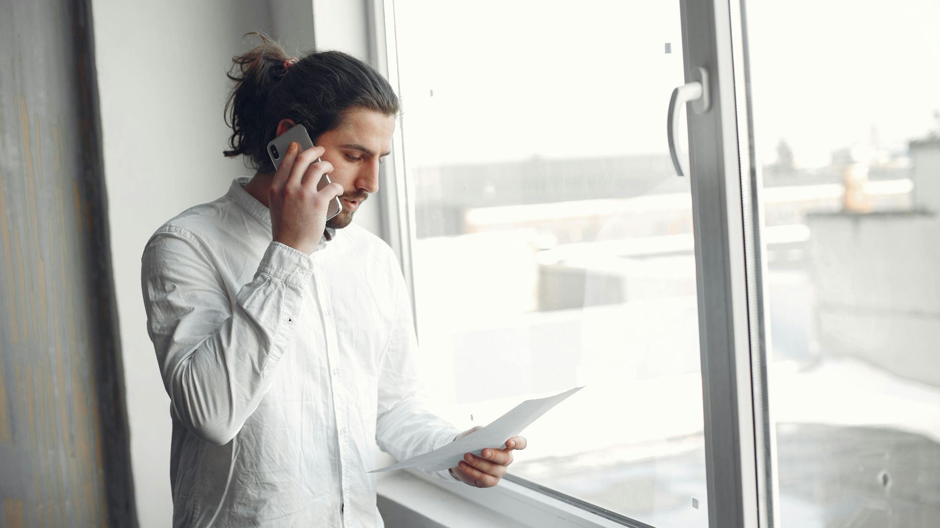 Young man in white shirt, on phone call holding a document, standing by a large window.