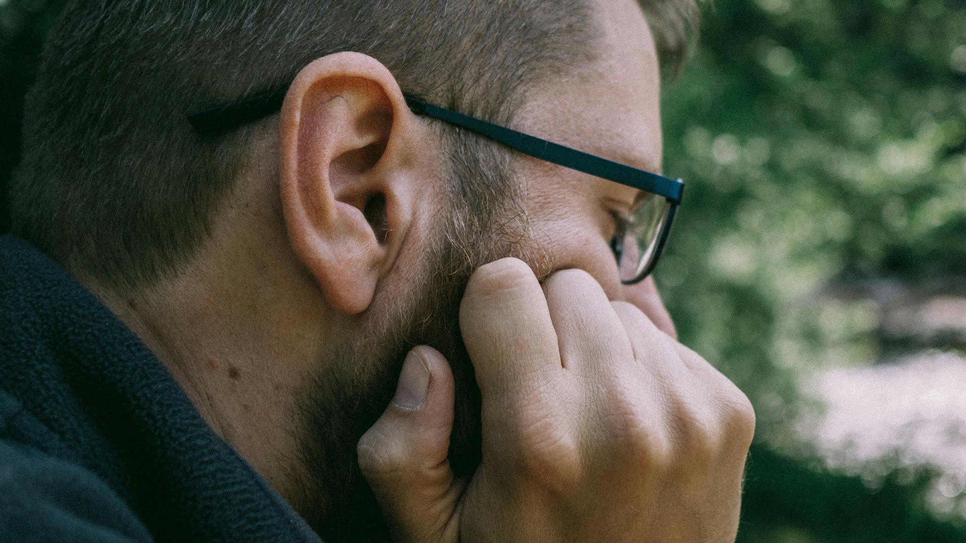A man with glasses contemplates while seated outdoors in lush greenery.