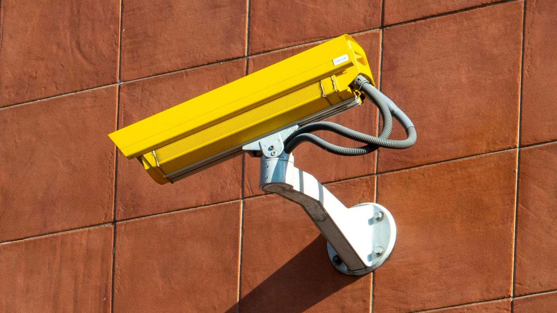 Security camera casting a shadow on an orange and brown tiled wall.
