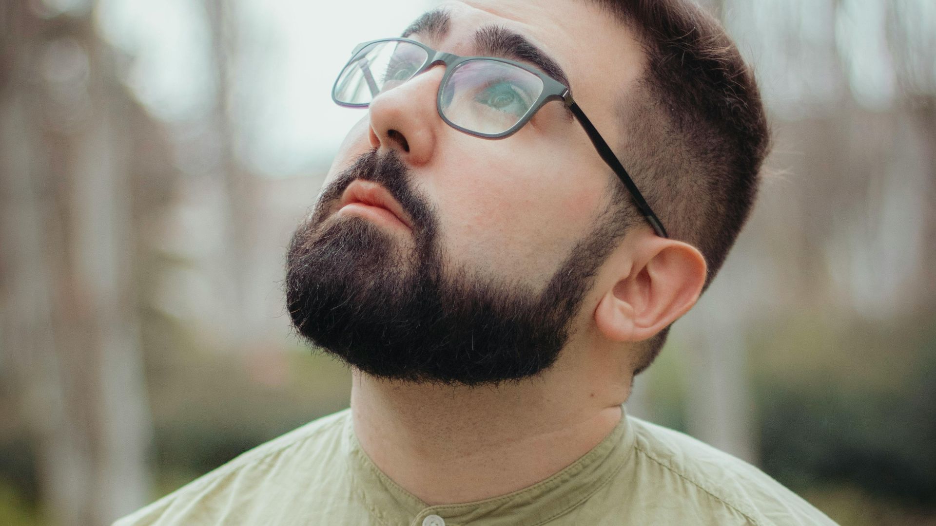 Portrait of a thoughtful man with beard and glasses, gazing upwards outdoors.
