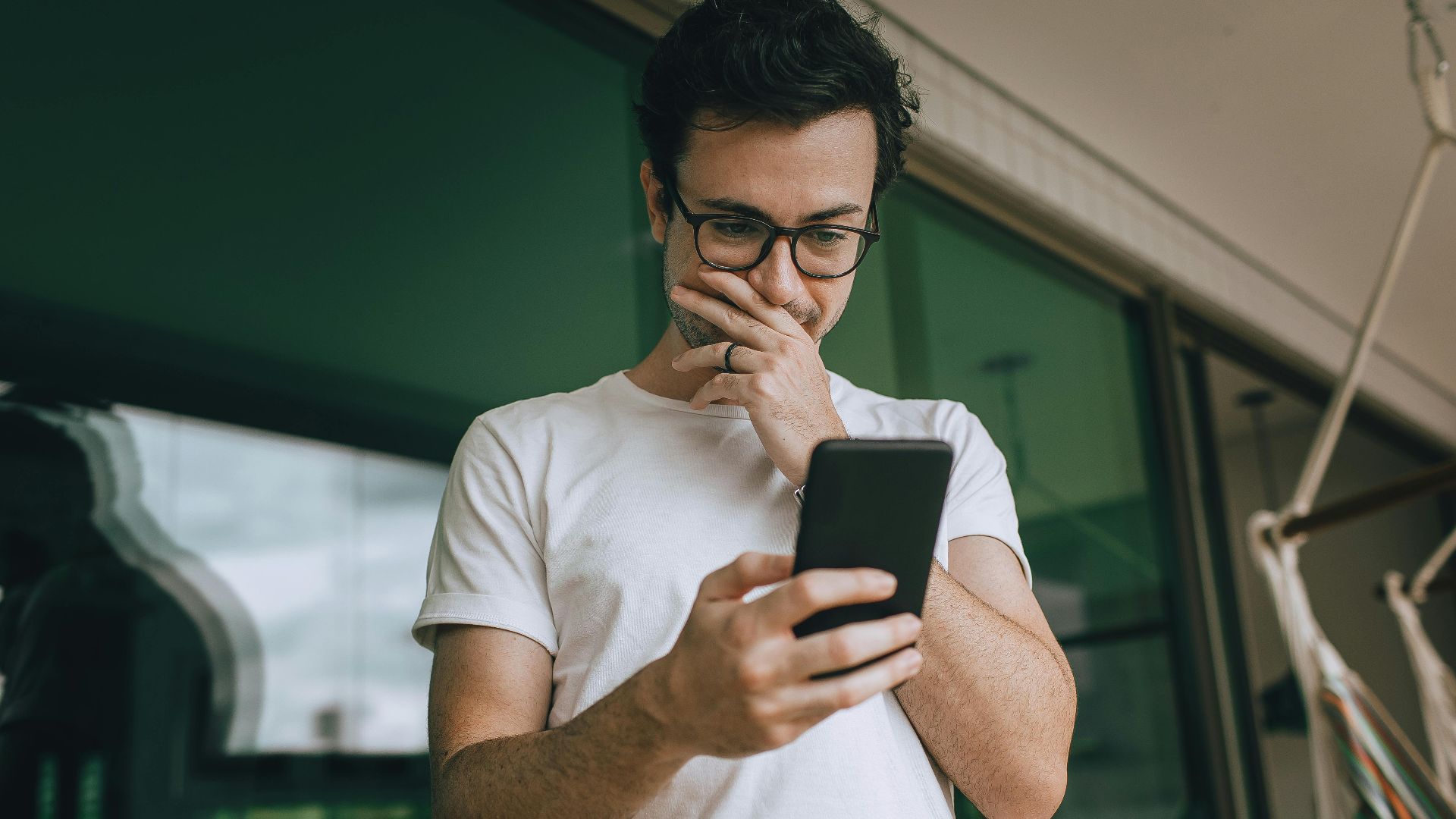 A young man in glasses standing on a balcony, using his smartphone with a thoughtful expression.