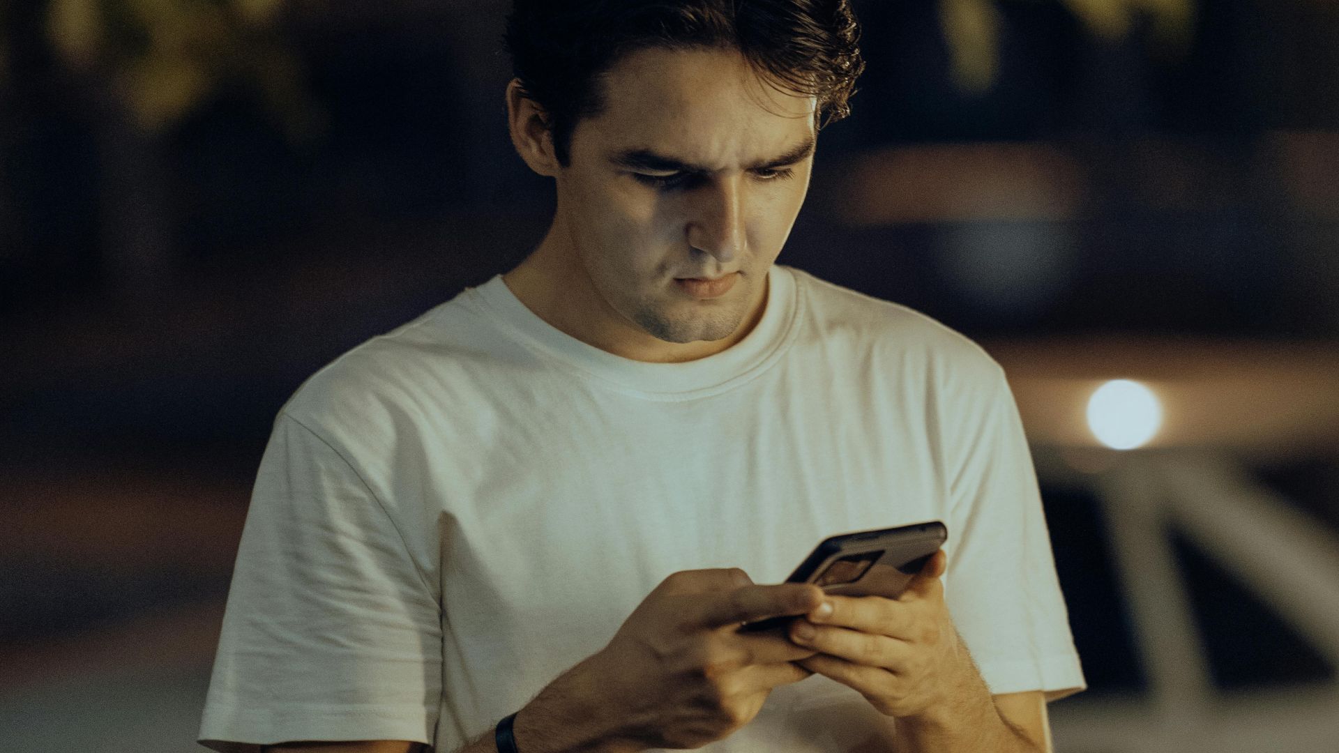 A young man in a white shirt stands outdoors using his smartphone during nighttime.