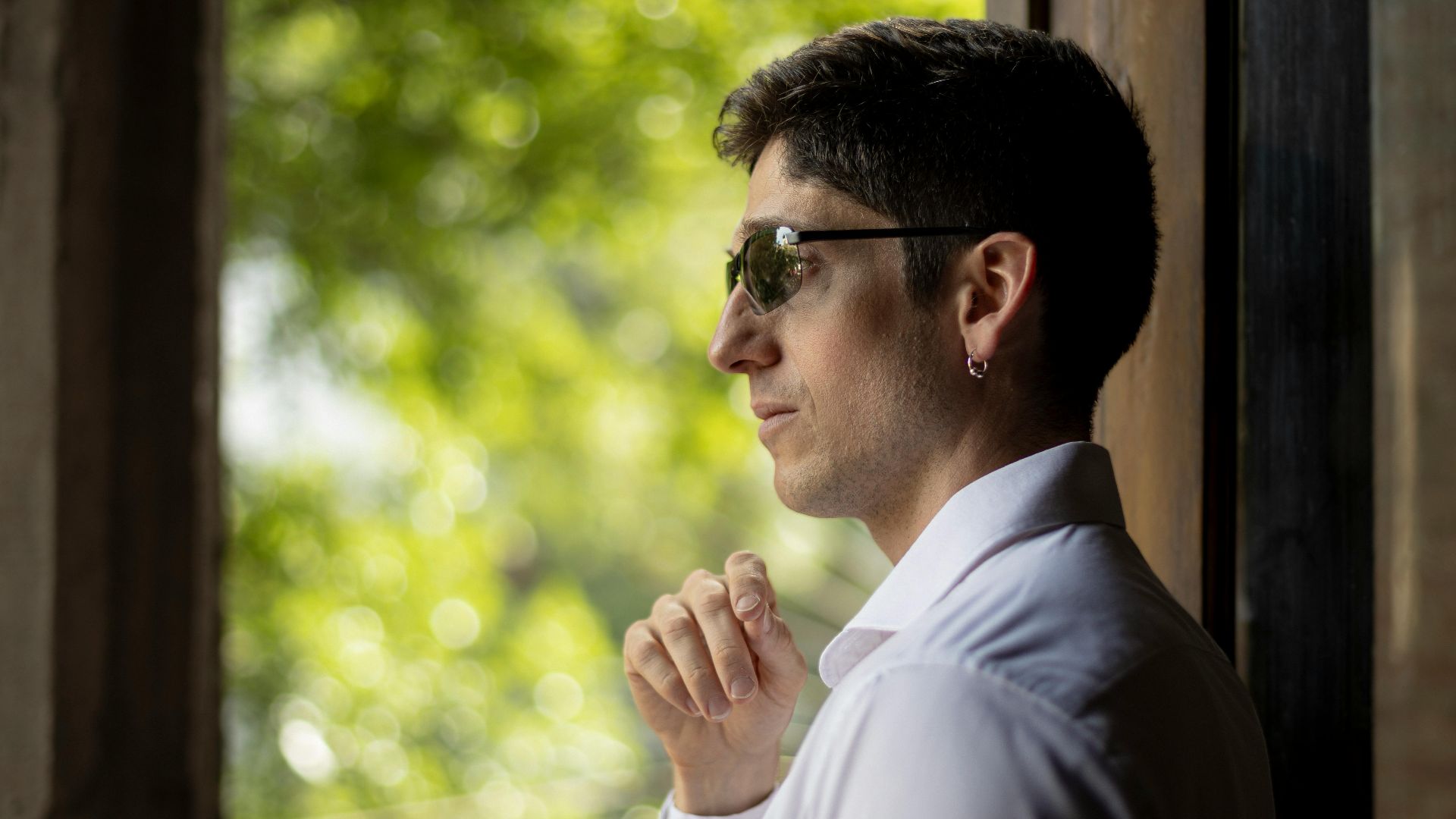 A stylish man wearing sunglasses gazes thoughtfully out a window, framed by greenery.