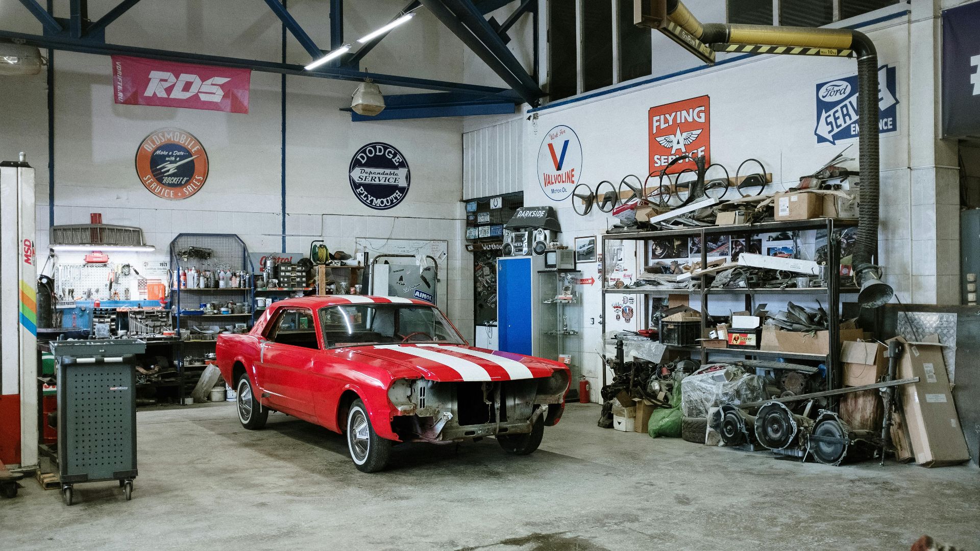 A vintage red car undergoing restoration in a well-equipped workshop with various tools and auto parts.