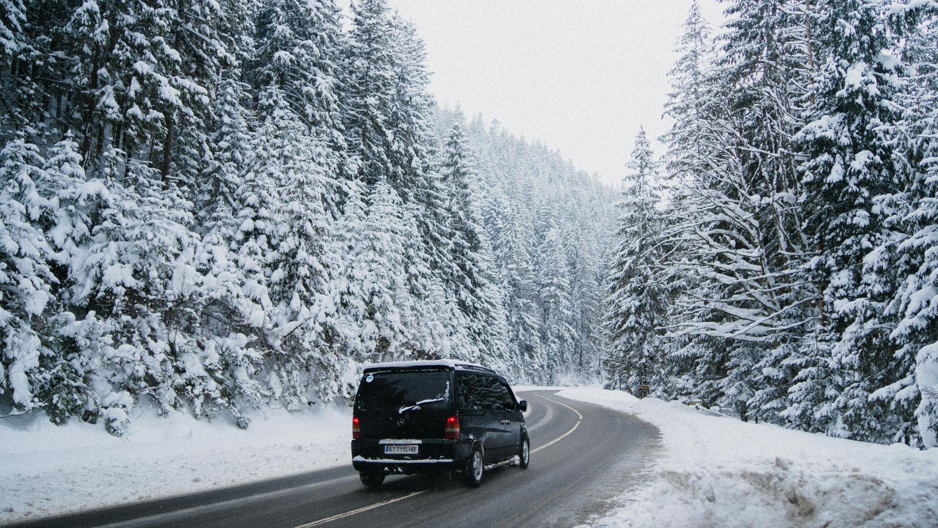 A black van driving on a snow-covered road through a winter forest with evergreen trees.