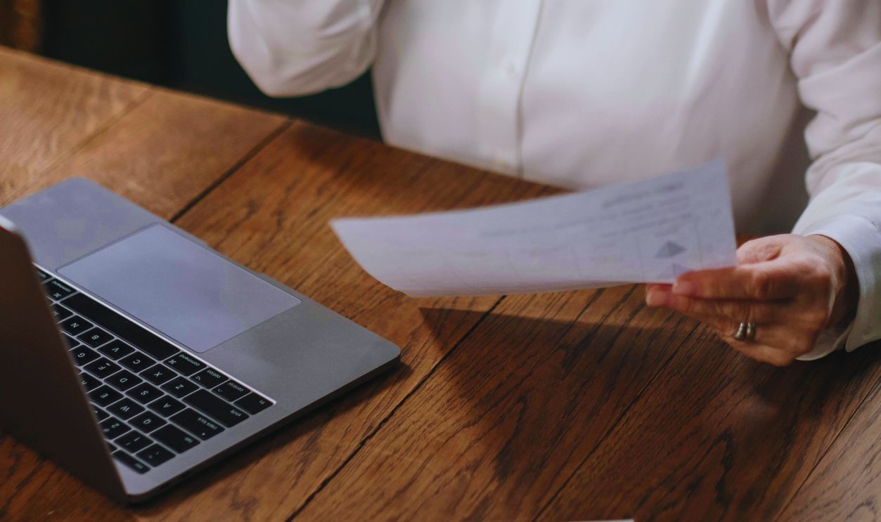 A Woman in White Dress Shirt Sitting at a Wooden Table Holding a Sheet of Paper