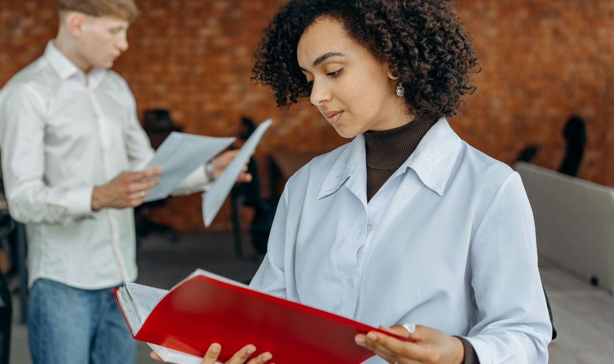 Employees Standing in White Long Sleeve Shirts Holding and Reading Paperwork Inside an Office