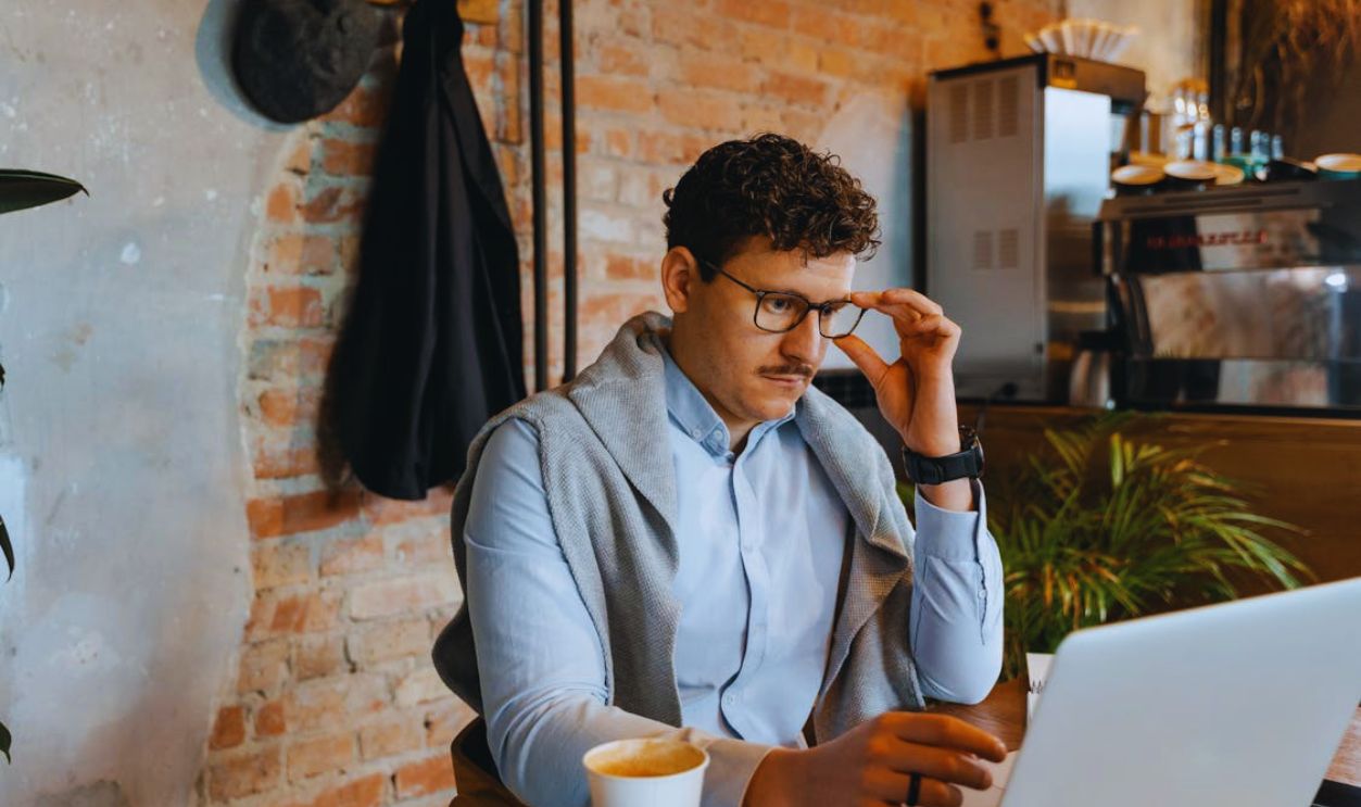 A Man Sitting at a Table with a Laptop Holding His Eyeglasses