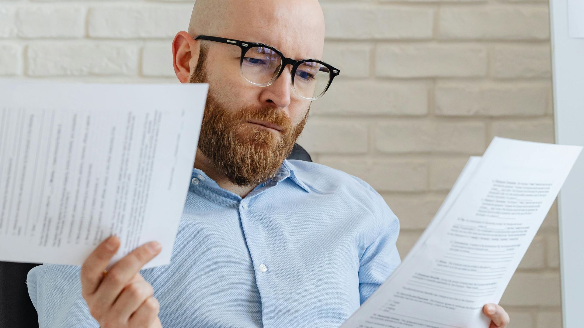 A serious-looking bald man with glasses reviews papers in an office against a brick wall.