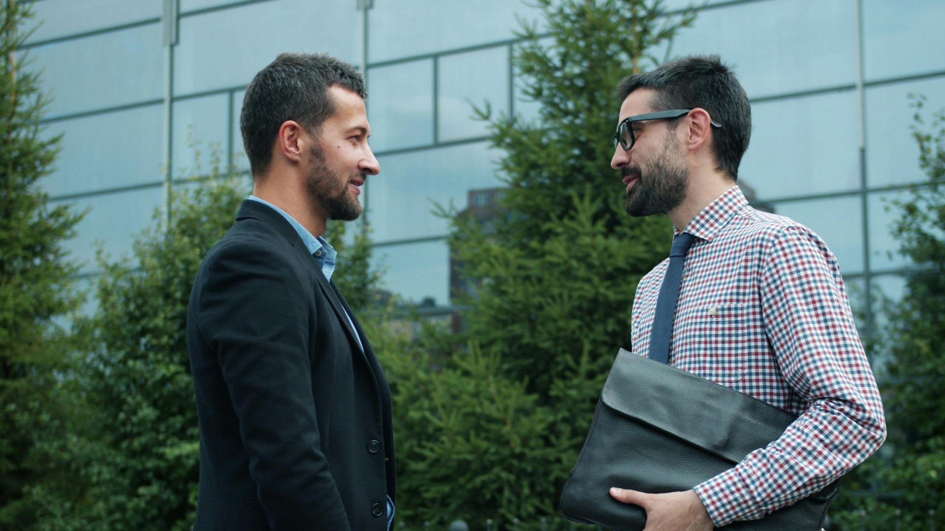 Two businessmen engage in conversation outdoors, standing beside a modern building.