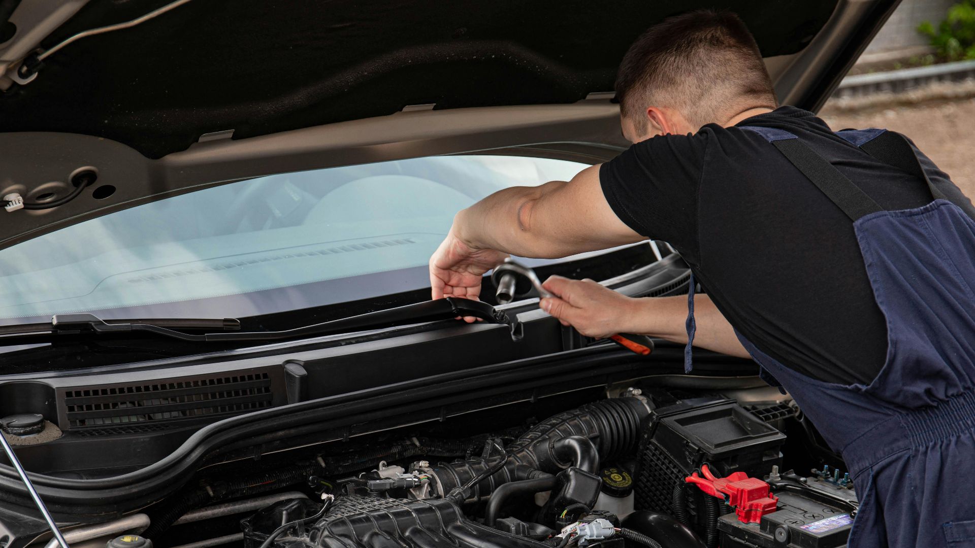 A mechanic uses tools to repair a car engine outdoors in daylight.