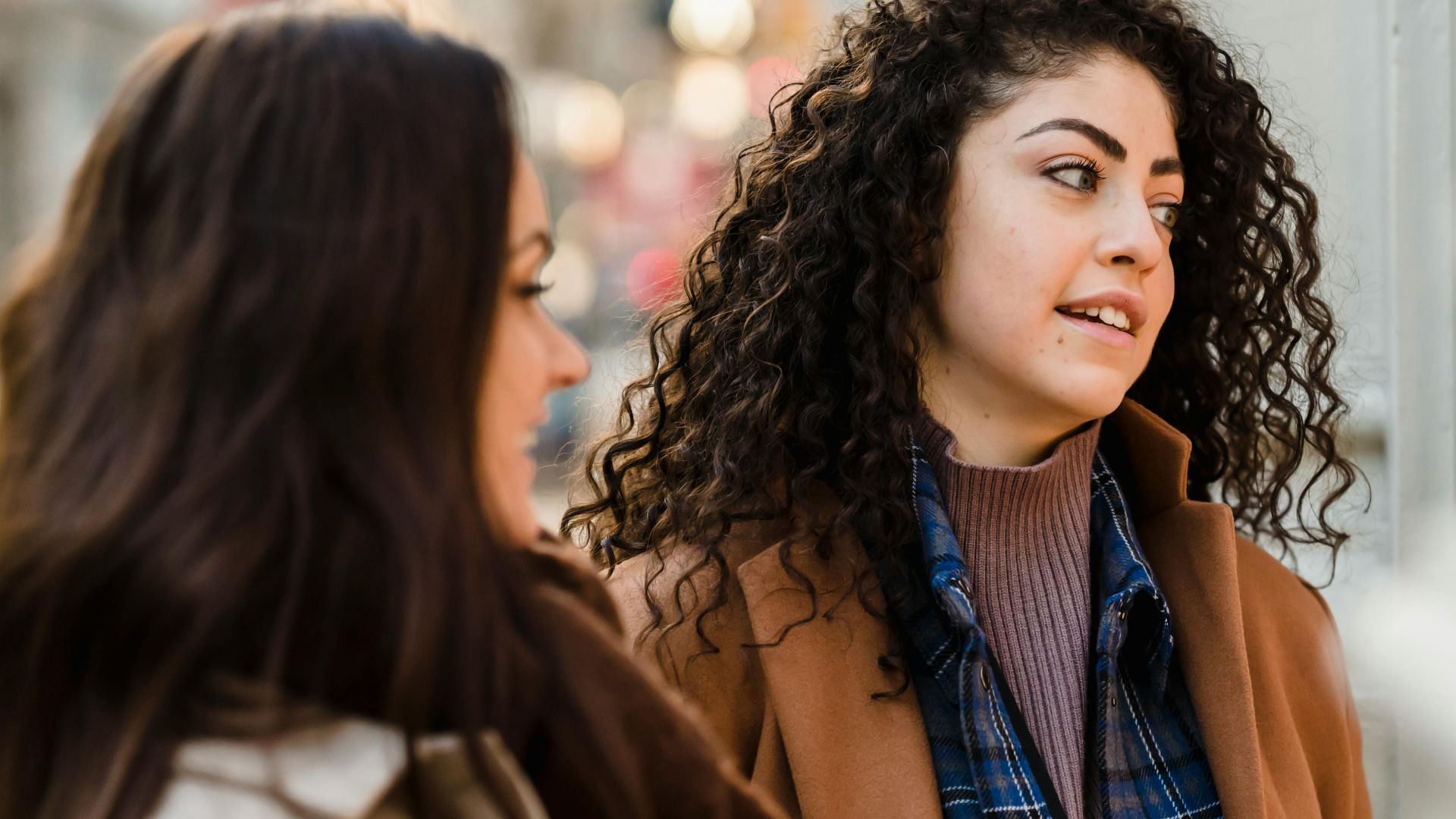 Optimistic young multiracial girlfriends with long dark hairs in warm clothes smiling while standing on street during weekend in city