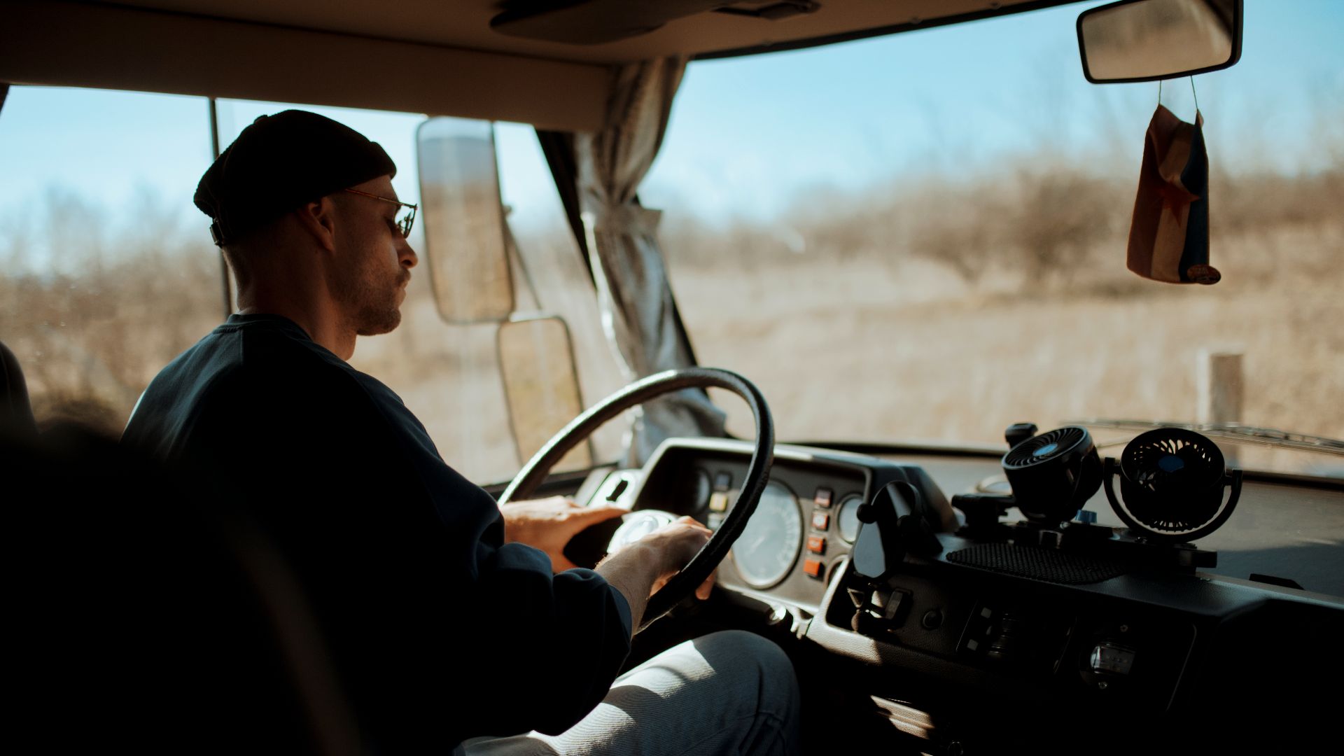 A person drives a truck on a clear day in an open area. The sun shines bright as the driver focuses on the road ahead. The landscape has trees in the background.