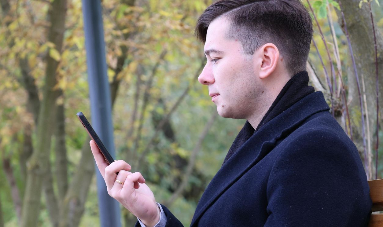 A Man in a Coat Sitting on a Wooden Bench Looking at His Smartphone