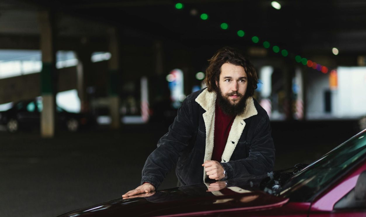 Young man poses with his car. Outdoor portrait of nice-looking positive guy in stylish outfit leaning on red car hood. Close-up handsome young man posing outdoors against modern shopping mall
