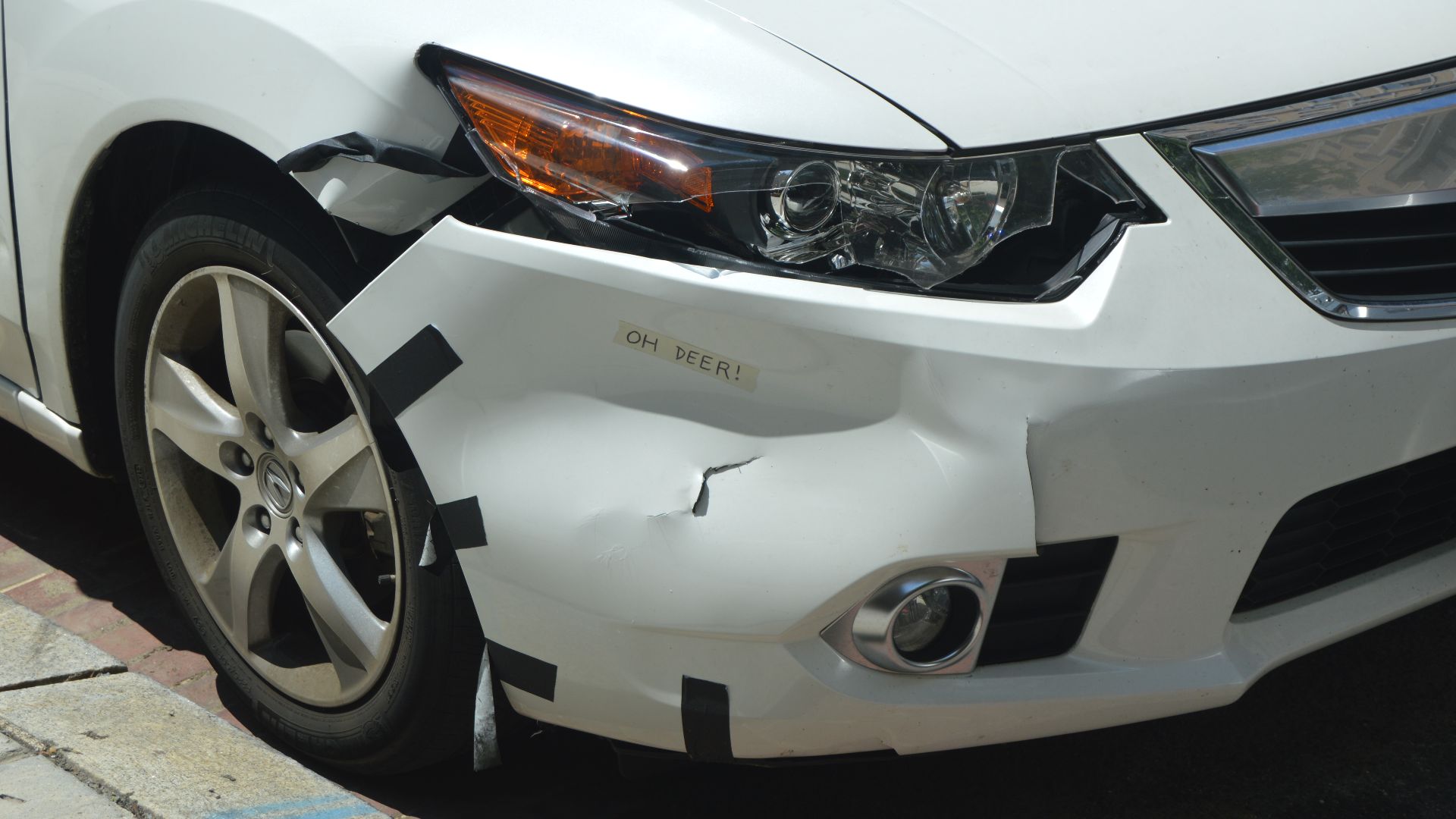 The results of a collision between a deer and an unidentified Acura car: the glass covering a headlight is smashed, much of the bumper and a small portion of the fender is cracked and impacted, and a piece of masking tape inscribed