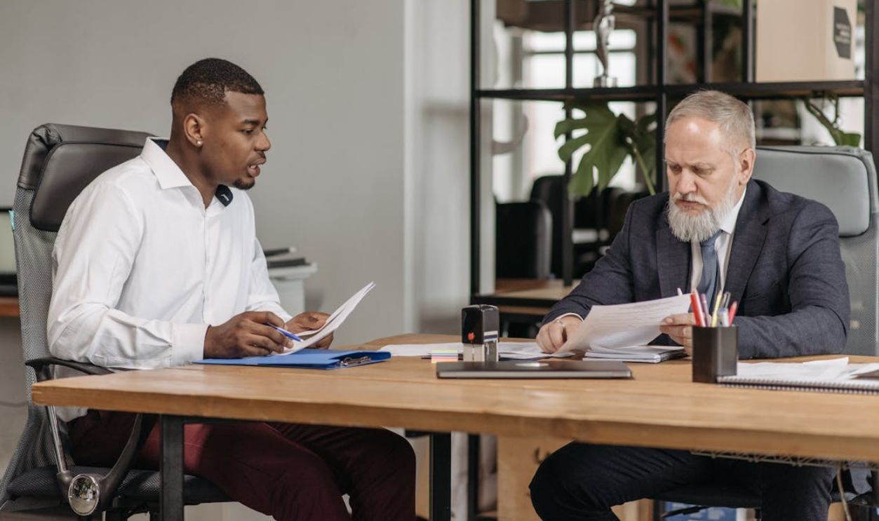 Elderly Man and Young Man Sitting at the Table in an Office and Looking at Documents