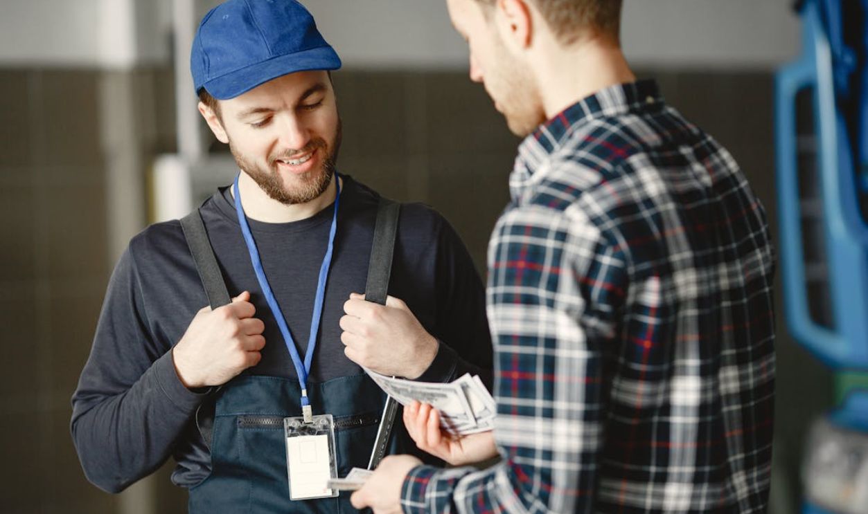Man in Plaid Shirt Giving Money to Bearded Man in Blue Cap and Overall