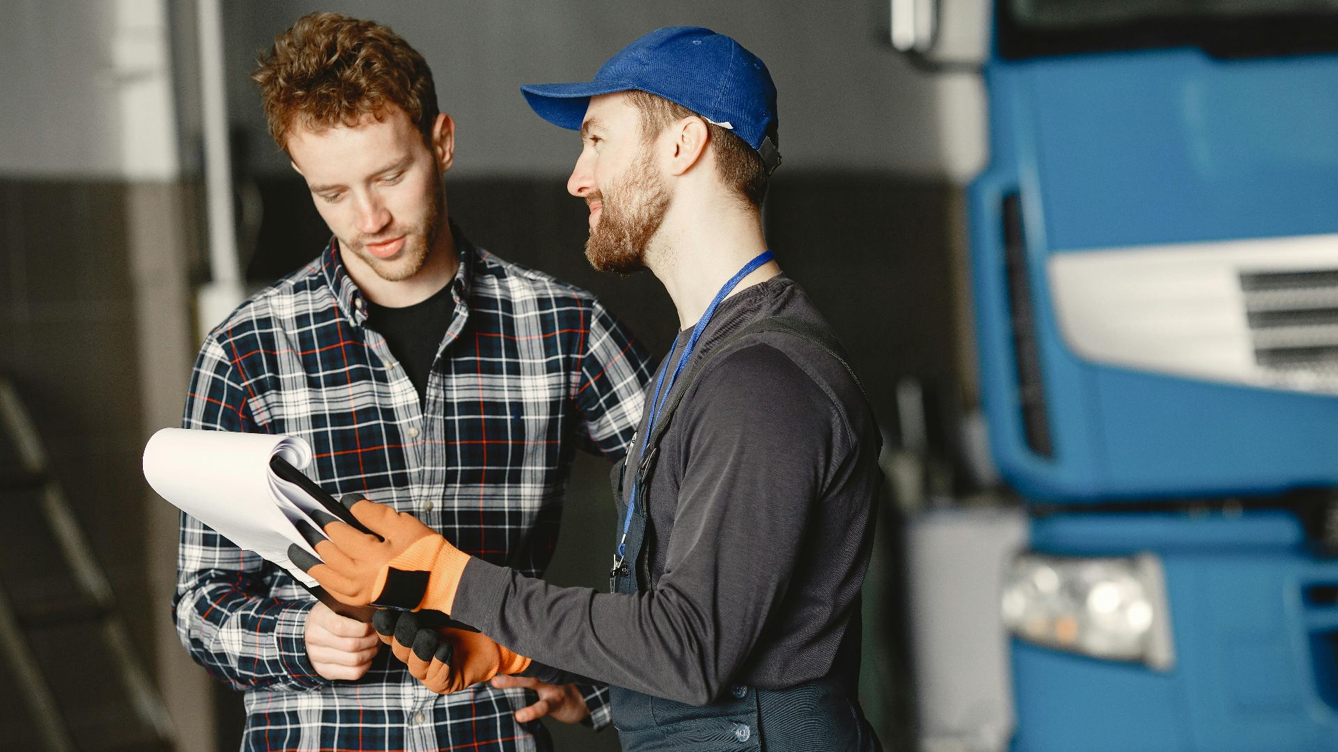 Mechanic in uniform consults with customer in garage setting beside a blue truck.