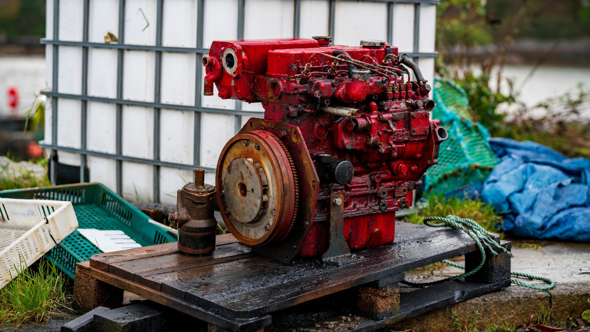 A rustic red diesel engine on a wooden pallet outdoors, surrounded by industrial objects.