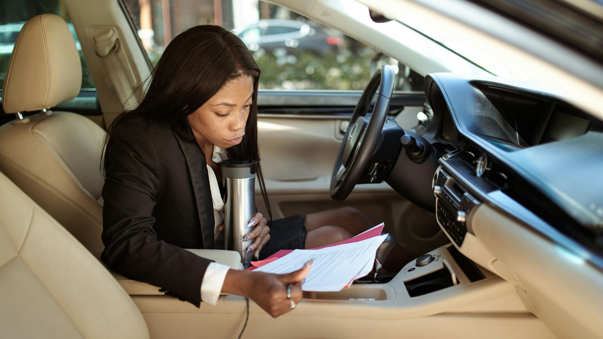 A focused businesswoman reads documents in her car, multitasking with a tumbler.