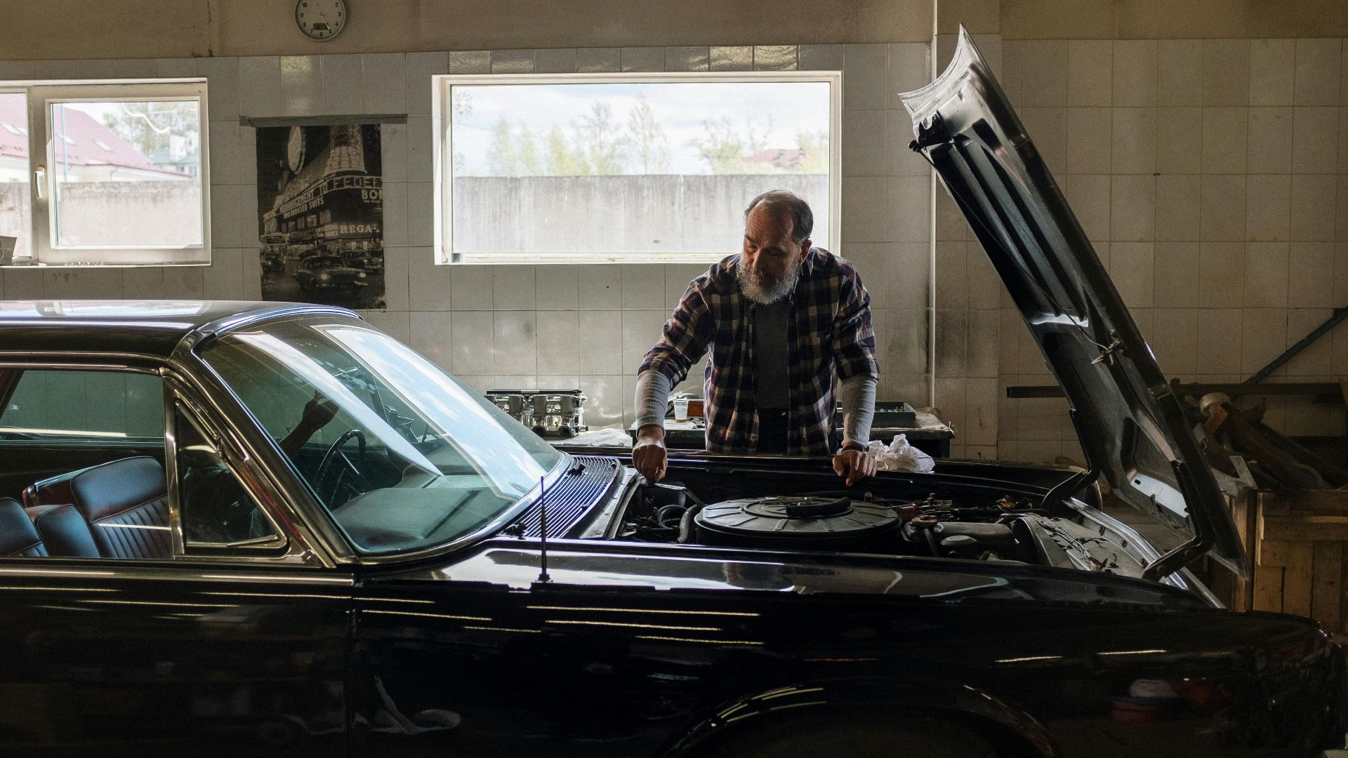 A bearded mechanic examines a classic car engine in a well-lit auto repair shop.