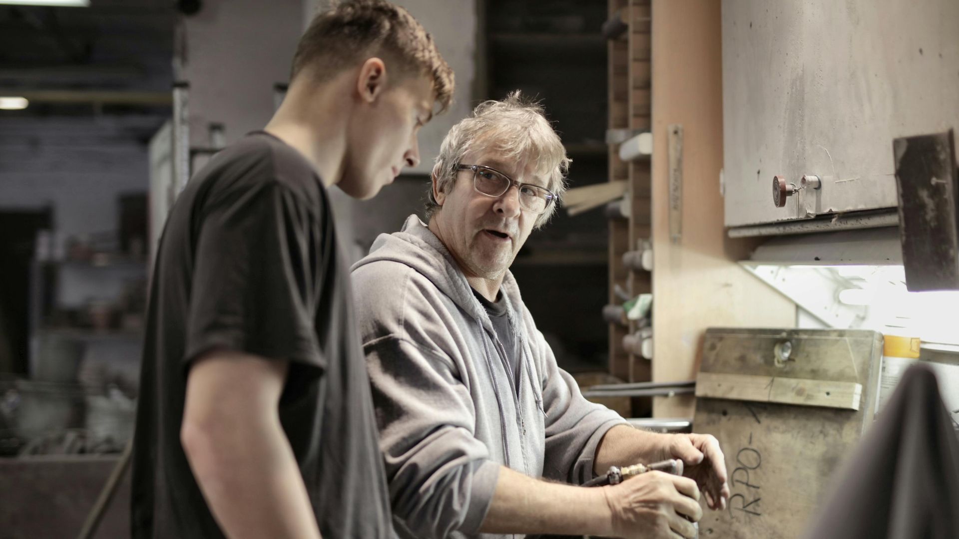 Side view of senior foreman in eyeglasses showing to trainee how handling detail in workshop