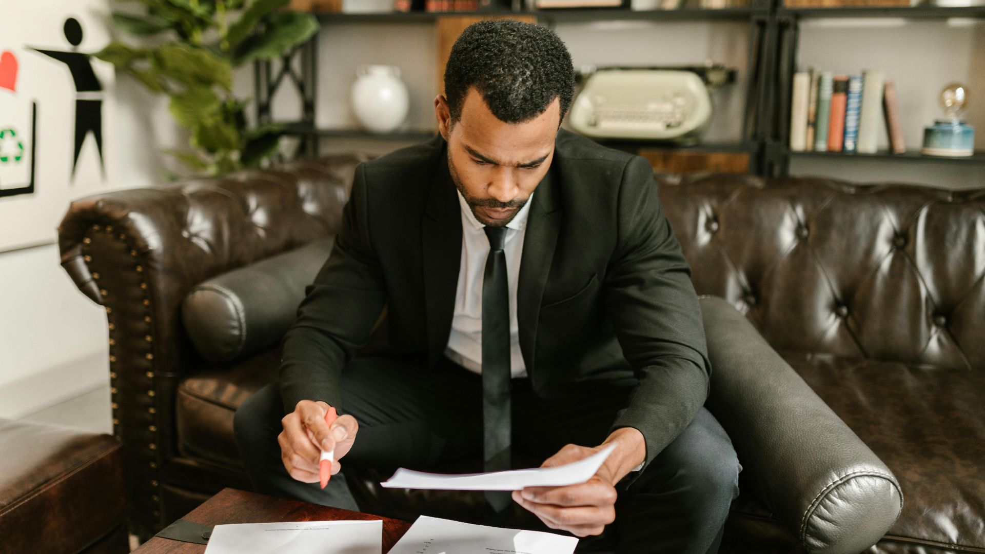 A businessman in a suit attentively reviews paperwork on a brown leather sofa.