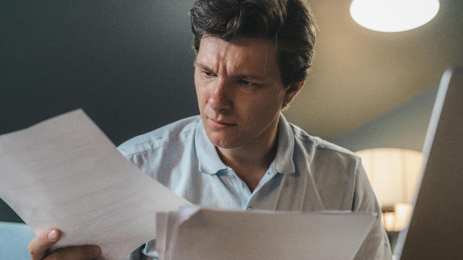 A man intently examines papers, seated indoors under warm lighting, focusing on his work.