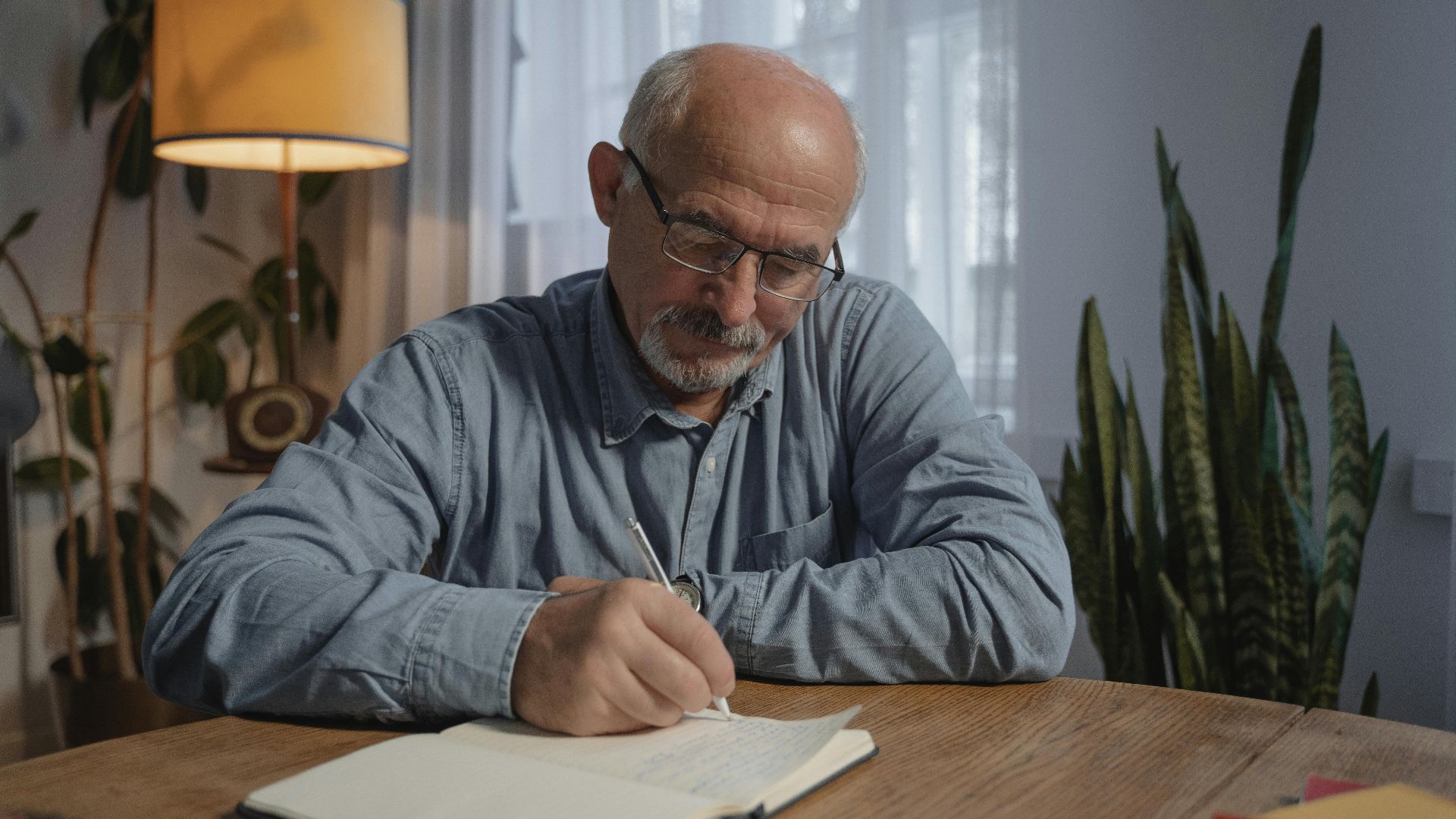 A senior adult man writing in a notebook at a desk in a cozy home office setting.