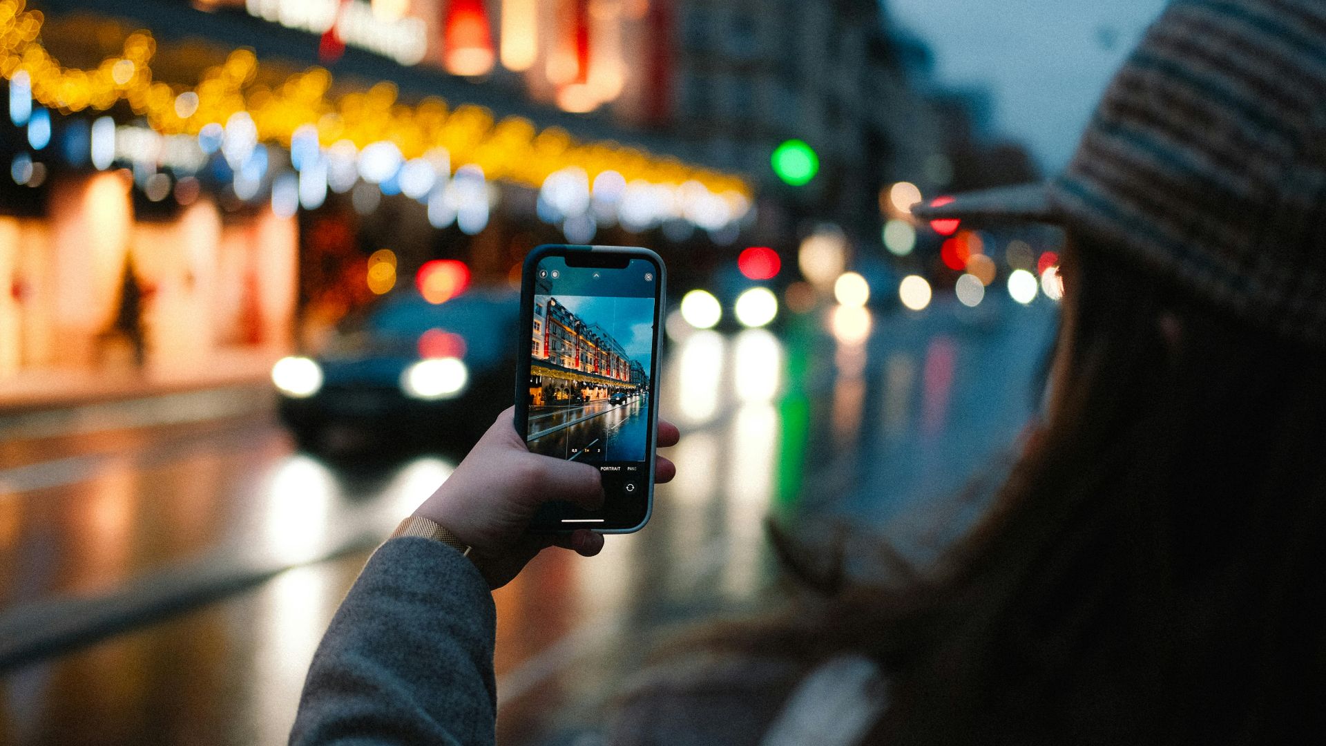 A woman photographs a vibrant city street with glowing lights and passing cars using her smartphone.