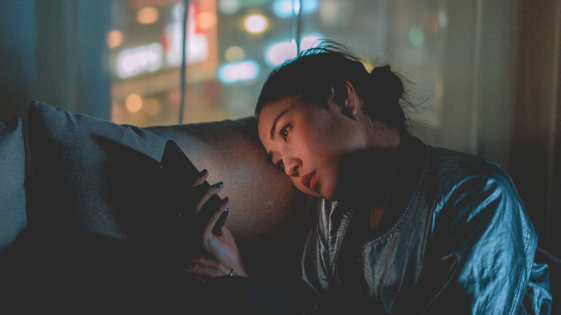 A woman looking at her mobile phone at night with city lights blurred in the background.