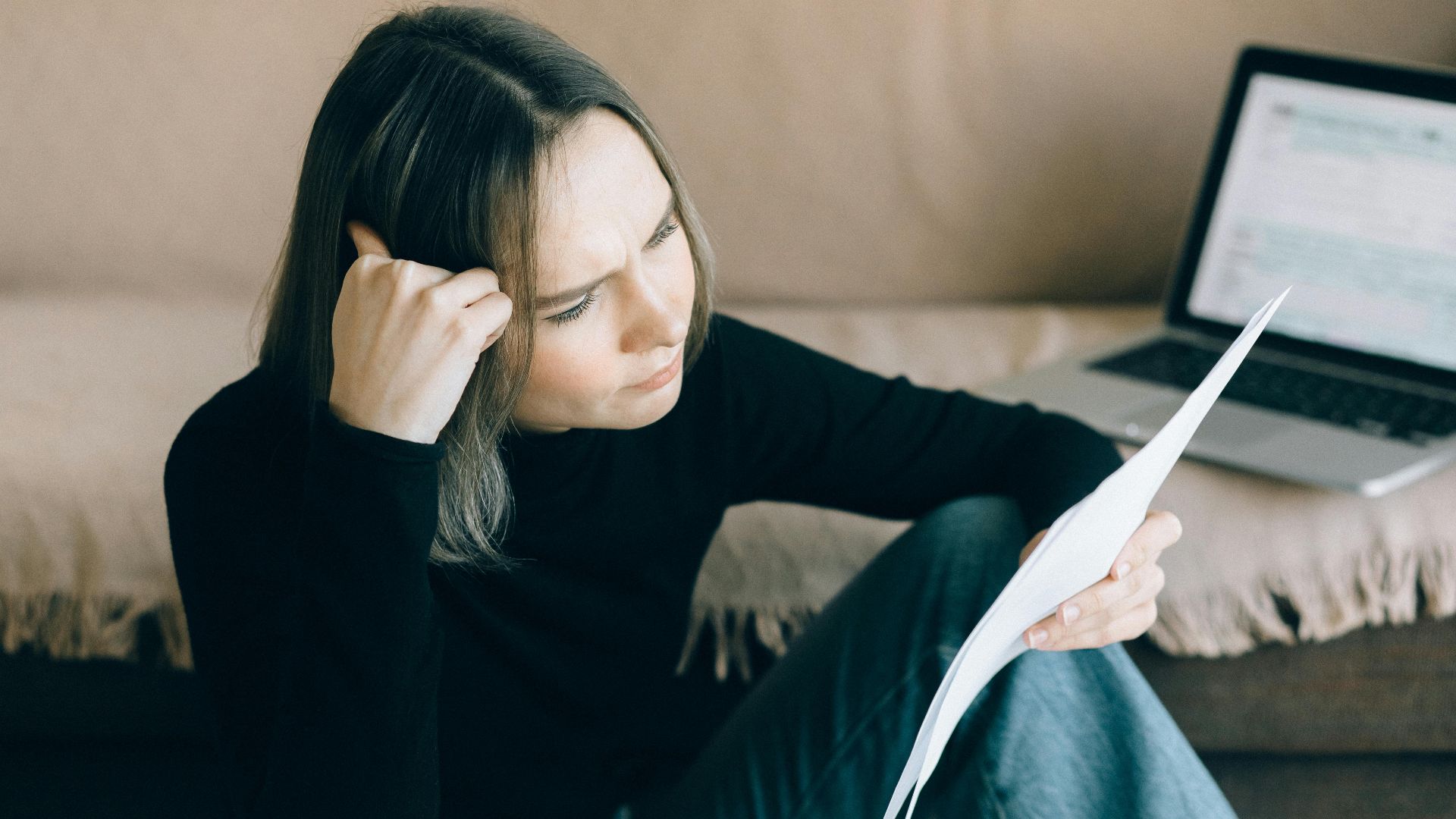 Focused woman analyzing papers with a laptop open, symbolizing thoughtful consideration.