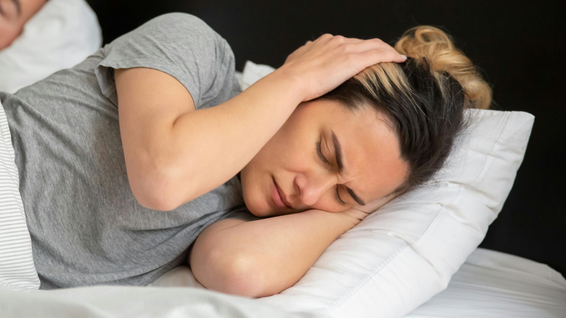 A woman looks frustrated in bed as her partner snores loudly, highlighting sleep disturbance issues.