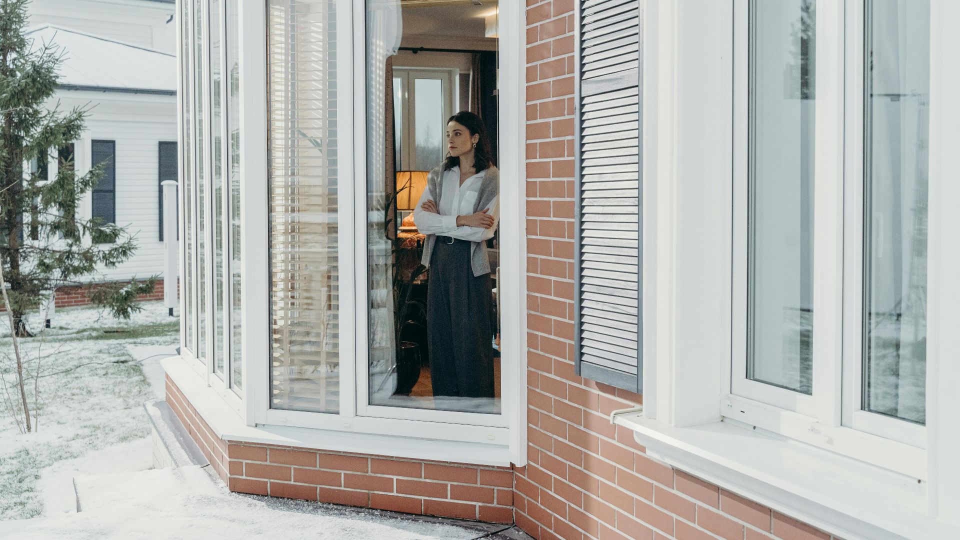 A woman stands indoors, arms crossed, looking out large windows on a snowy day, reflecting indoors and outdoors contrast.