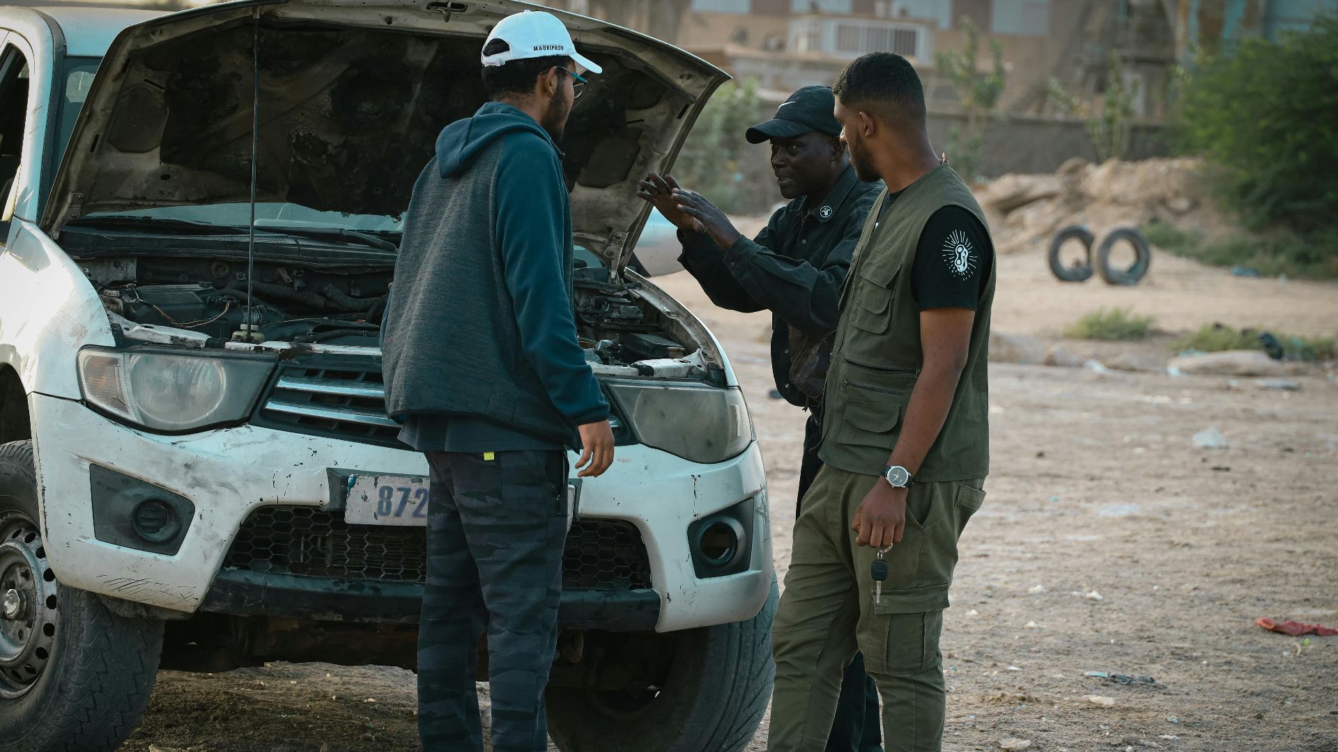Three men examine a car with an open hood in an outdoor setting, discussing possible repairs.