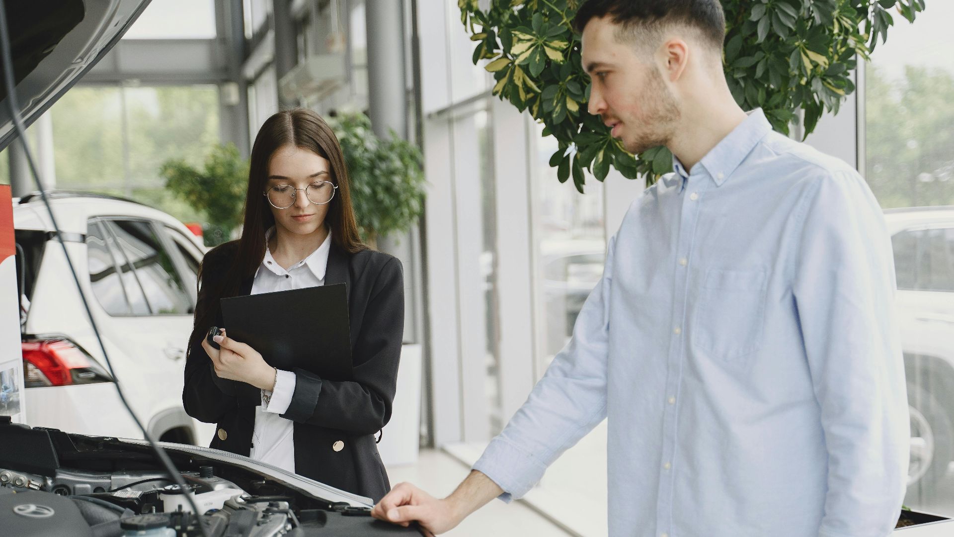 Man and woman examining car engine in dealership service area, focused on vehicle maintenance and functionality.