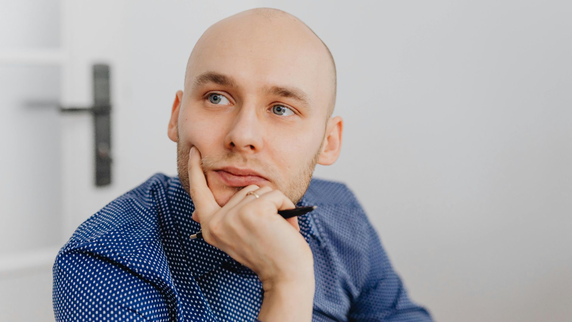 Bald man in blue shirt thoughtfully holding a pen indoors, gazing into distance.