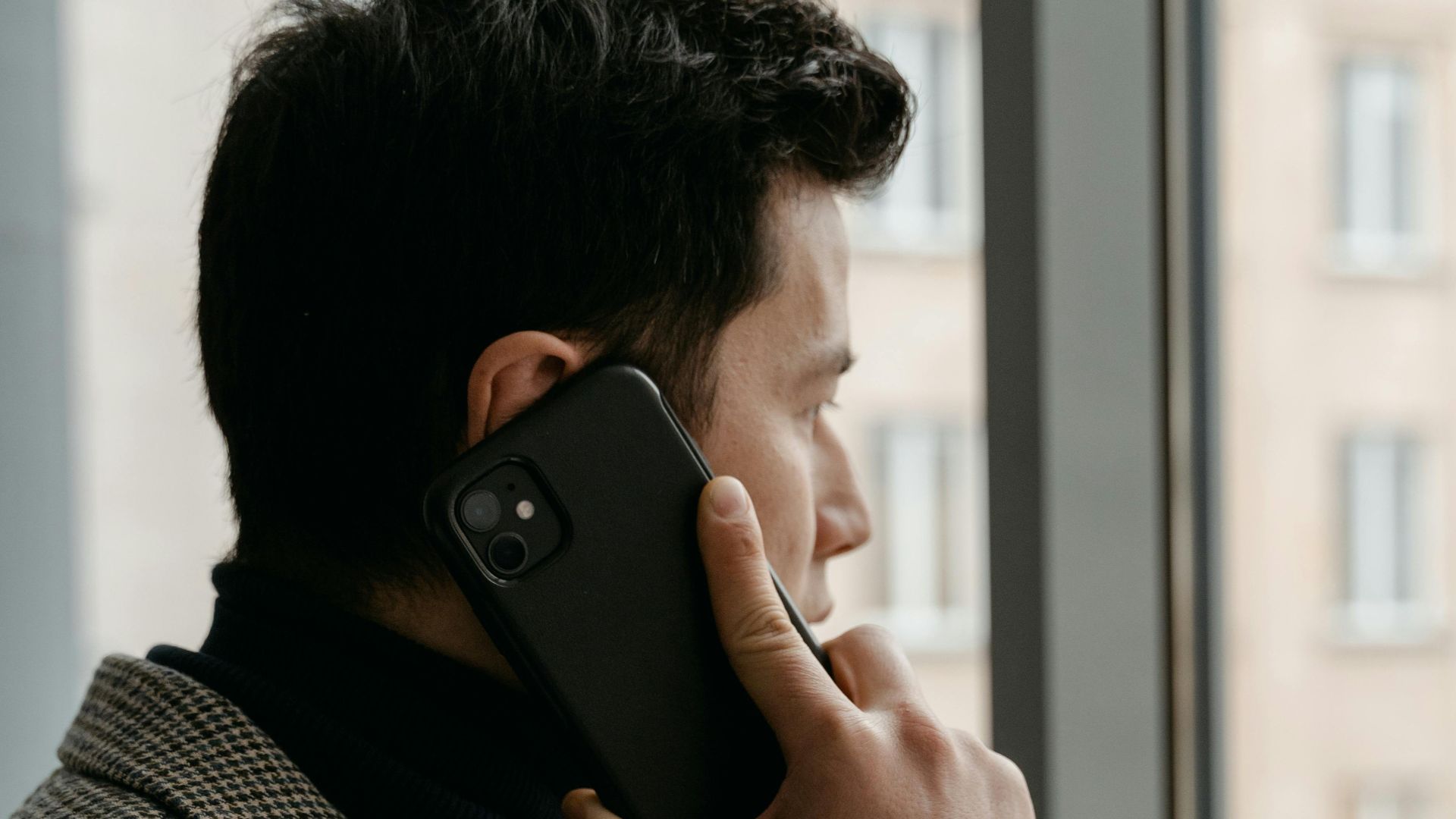 Man in a checkered blazer using a smartphone while gazing out a window.
