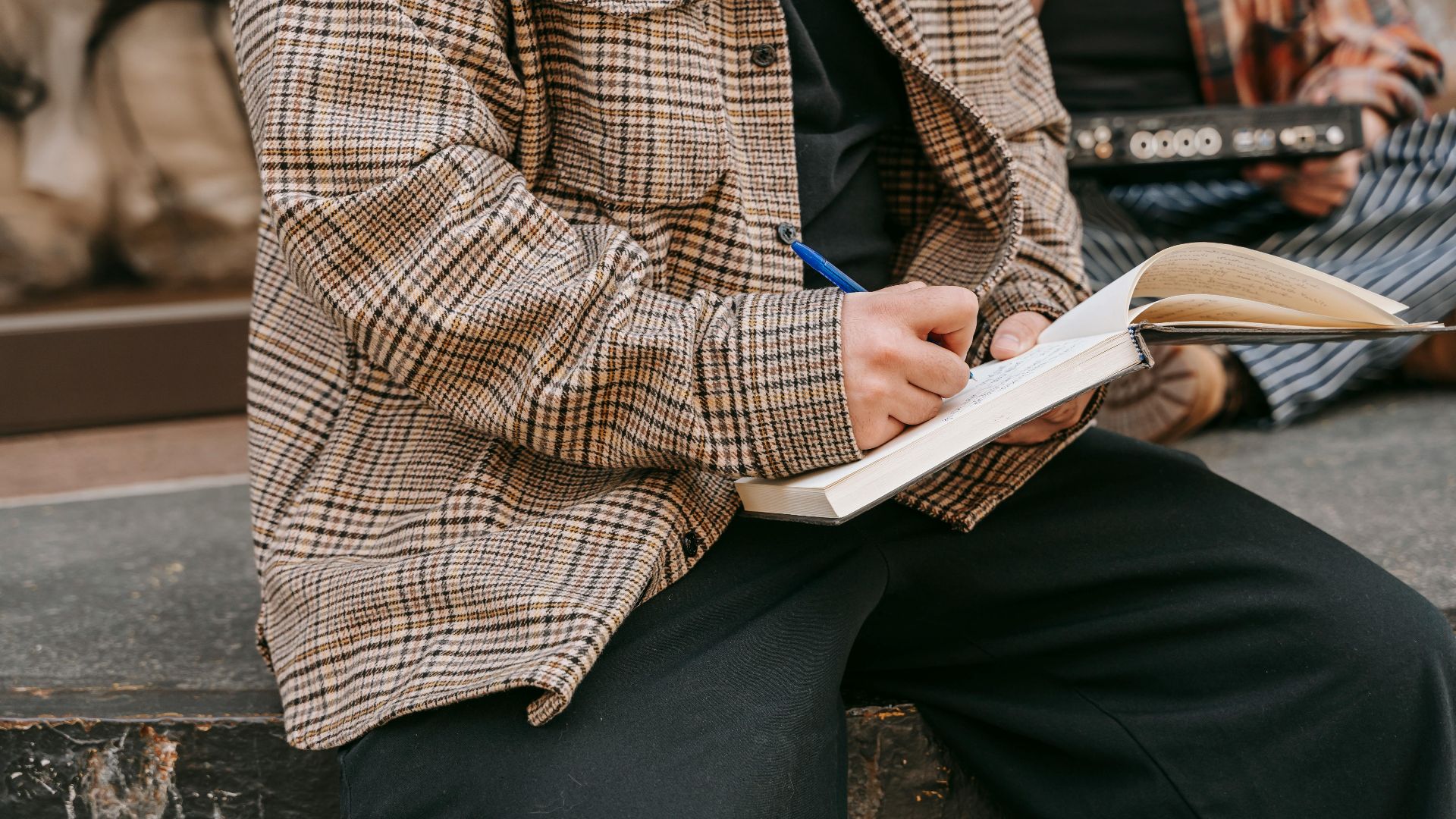 Close-up of a person writing in a notebook with a pen, sitting outdoors on a sunny day.