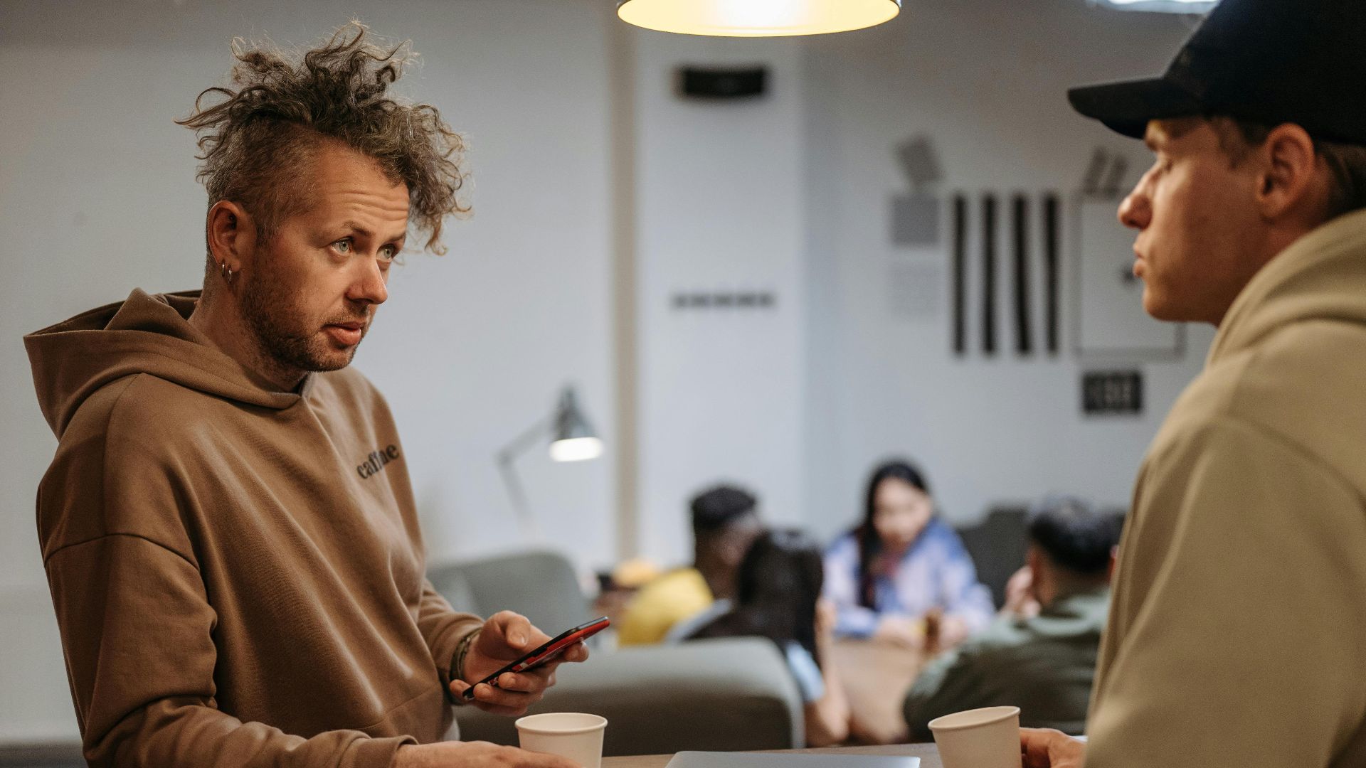 Two men in a casual office setting talking over coffee with laptops.