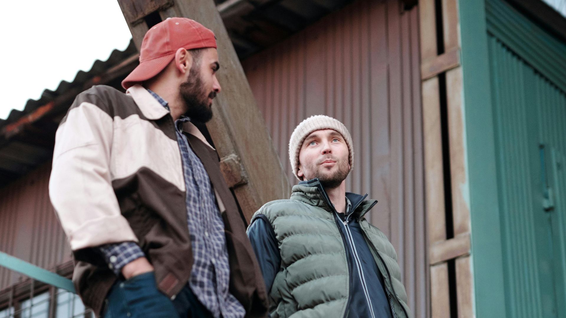 Two men engage in conversation on industrial metal staircase, wearing casual winter clothing.