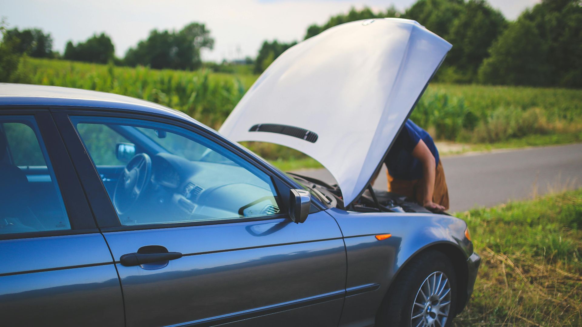 Man checking car engine with hood open by the side of a rural road.