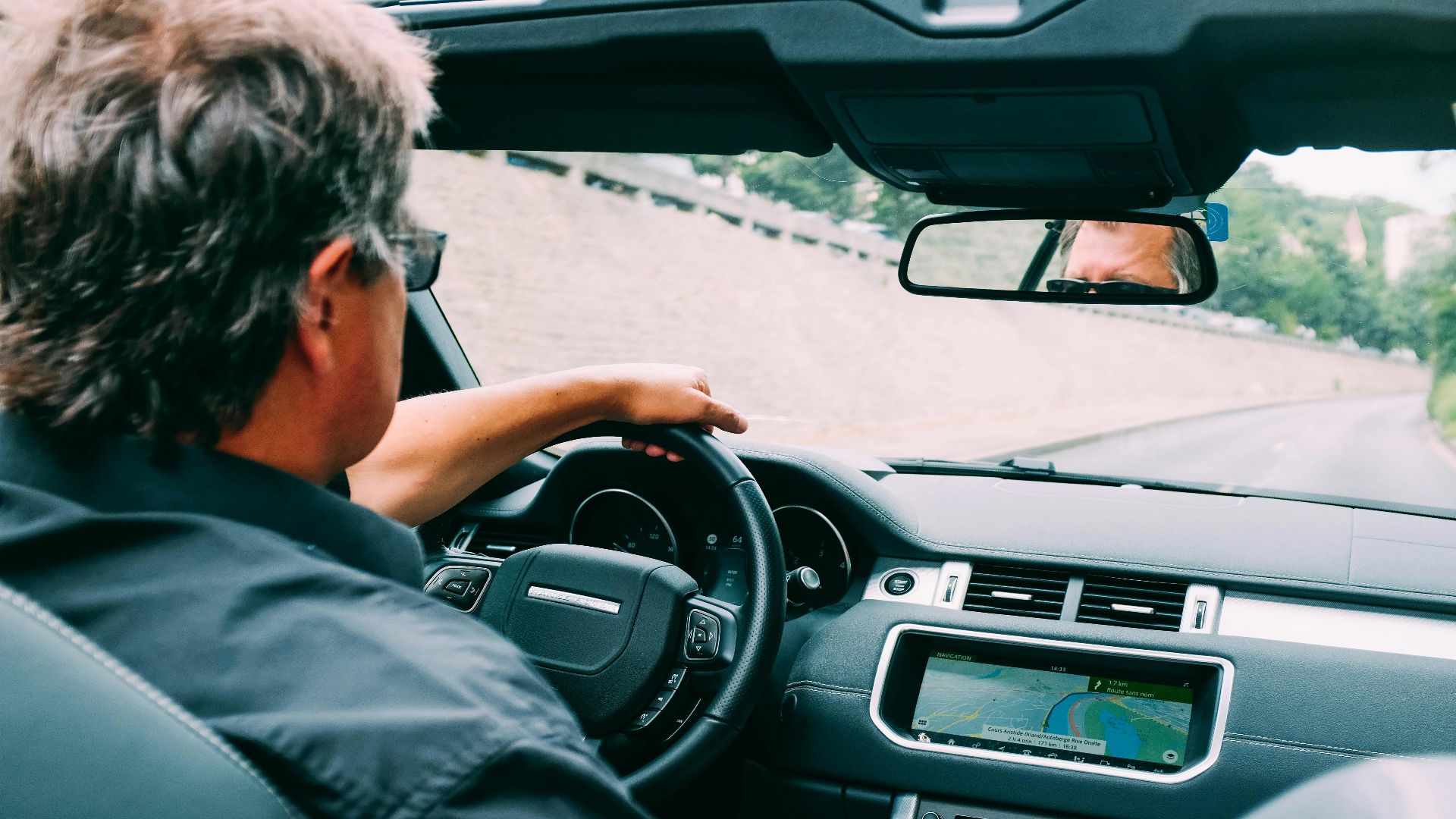 A man driving a convertible car on a scenic highway with a focus on the dashboard.