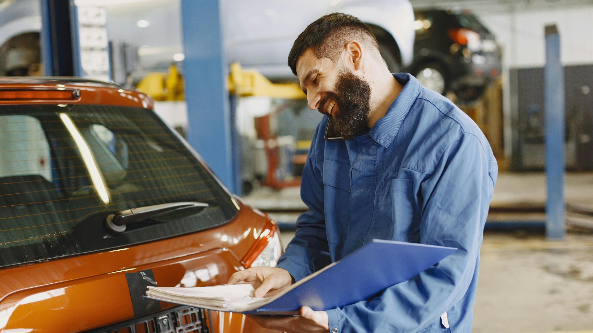 Smiling auto mechanic inspects a car in a service garage, wearing coveralls and holding documents.