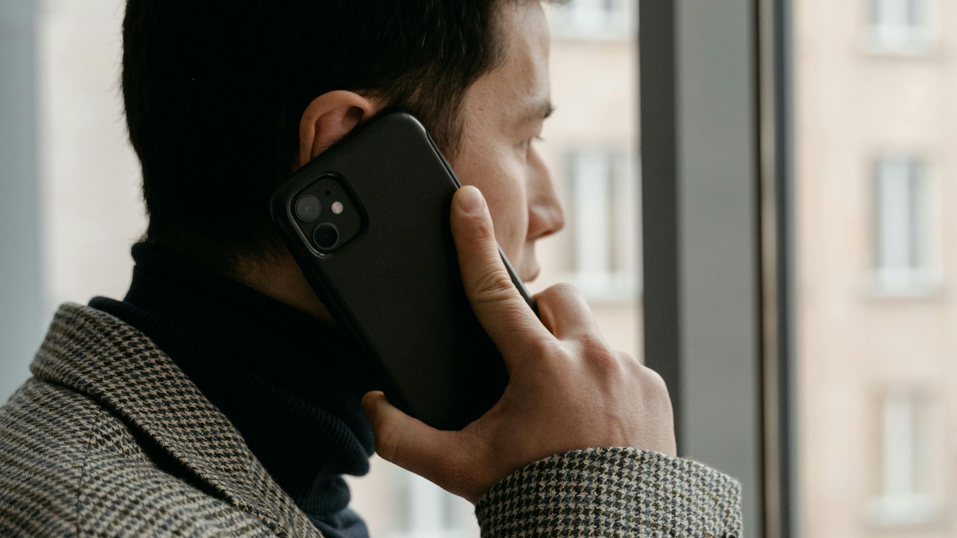 Man in a checkered blazer using a smartphone while gazing out a window.