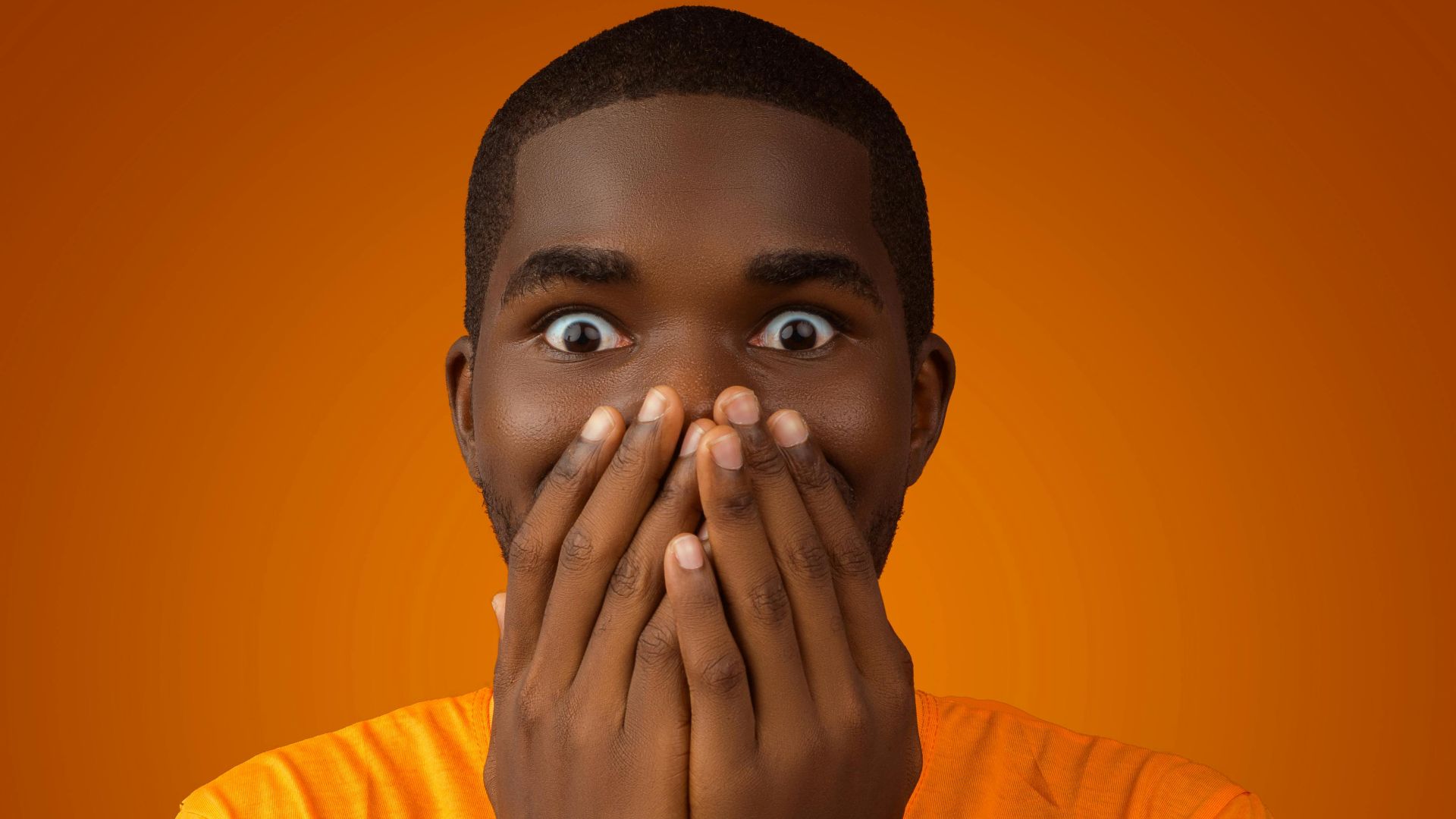 A surprised man with wide eyes covering his mouth in shock against an orange background.