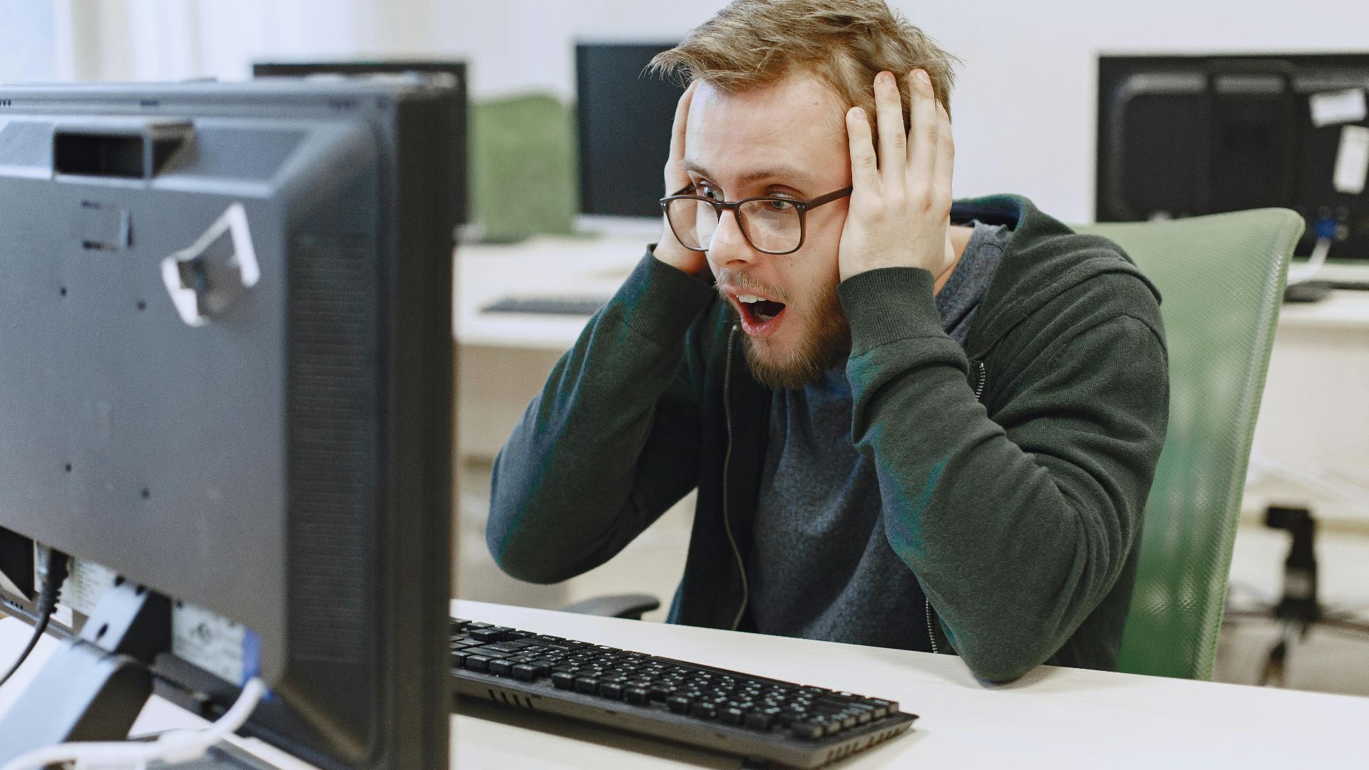 Surprised man in hoodie reacting to shocking news on computer monitor while sitting in a modern office.