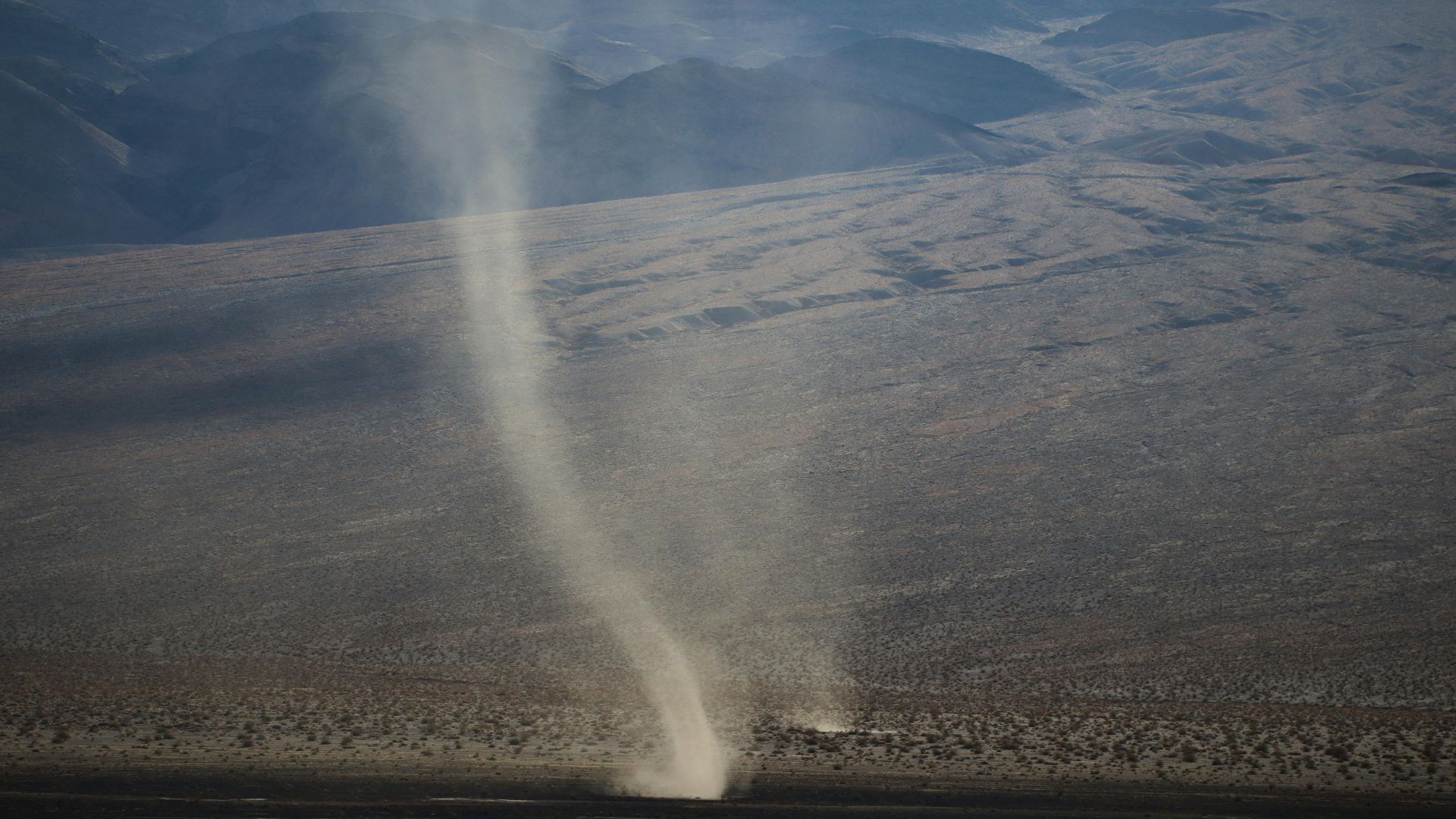 Aerial view of a dramatic dust devil swirling in a vast arid desert landscape under clear skies.